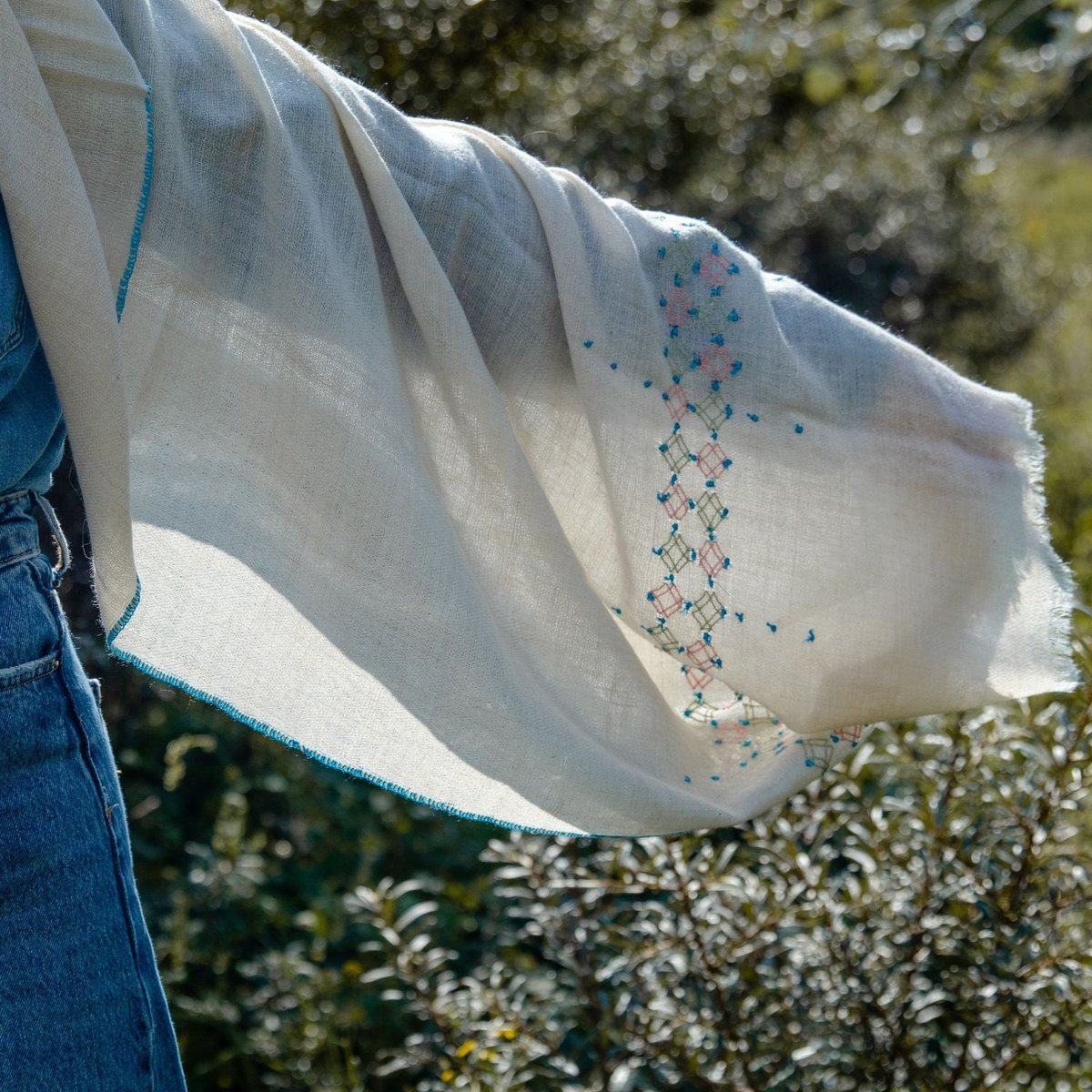 A cream pashmina held out in the wind, cross-stitch diamonds in teal, pink and olive along it