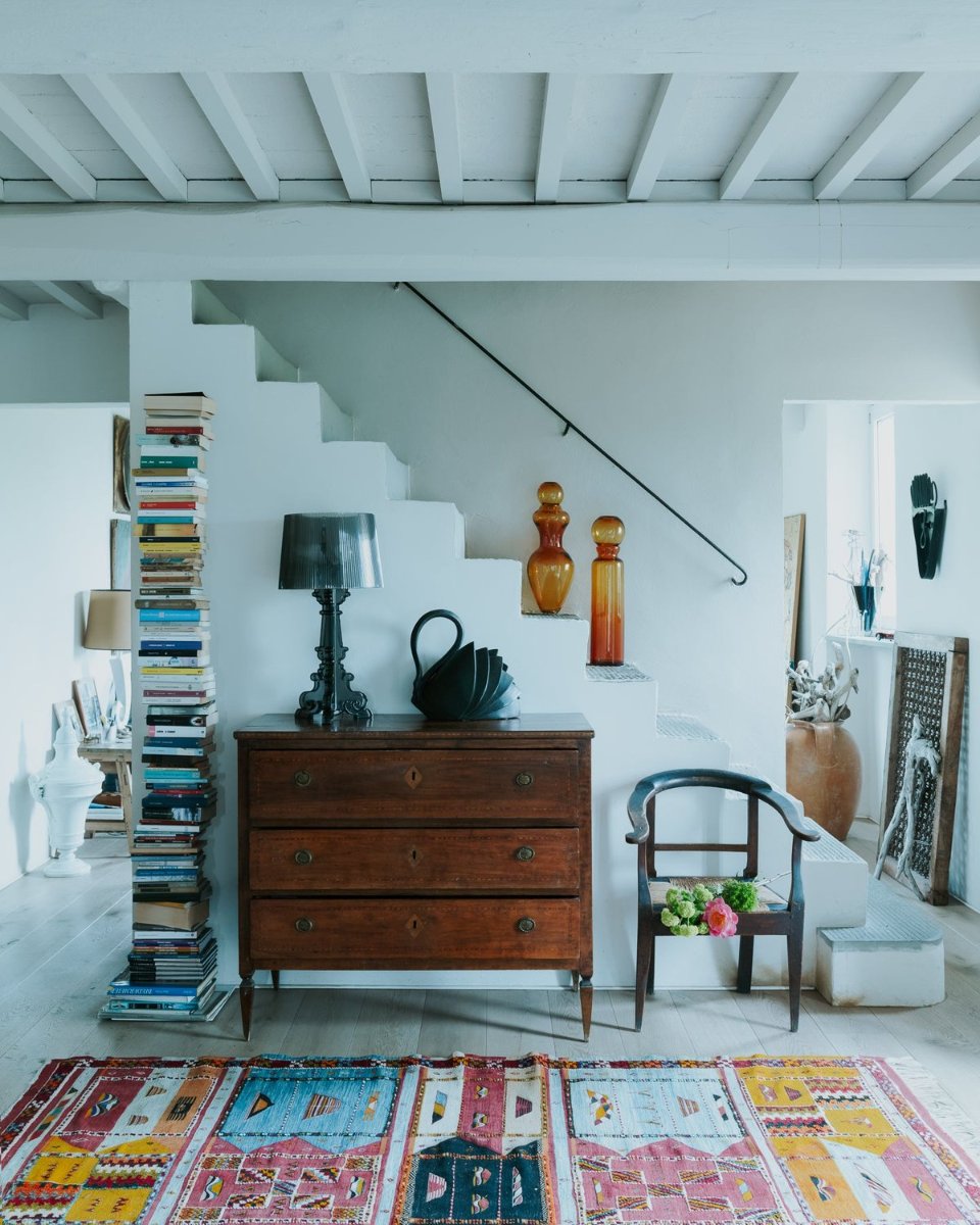A whitewashed room with beamed ceilings, a dark chest below the stairs, two amber bottles on the steps