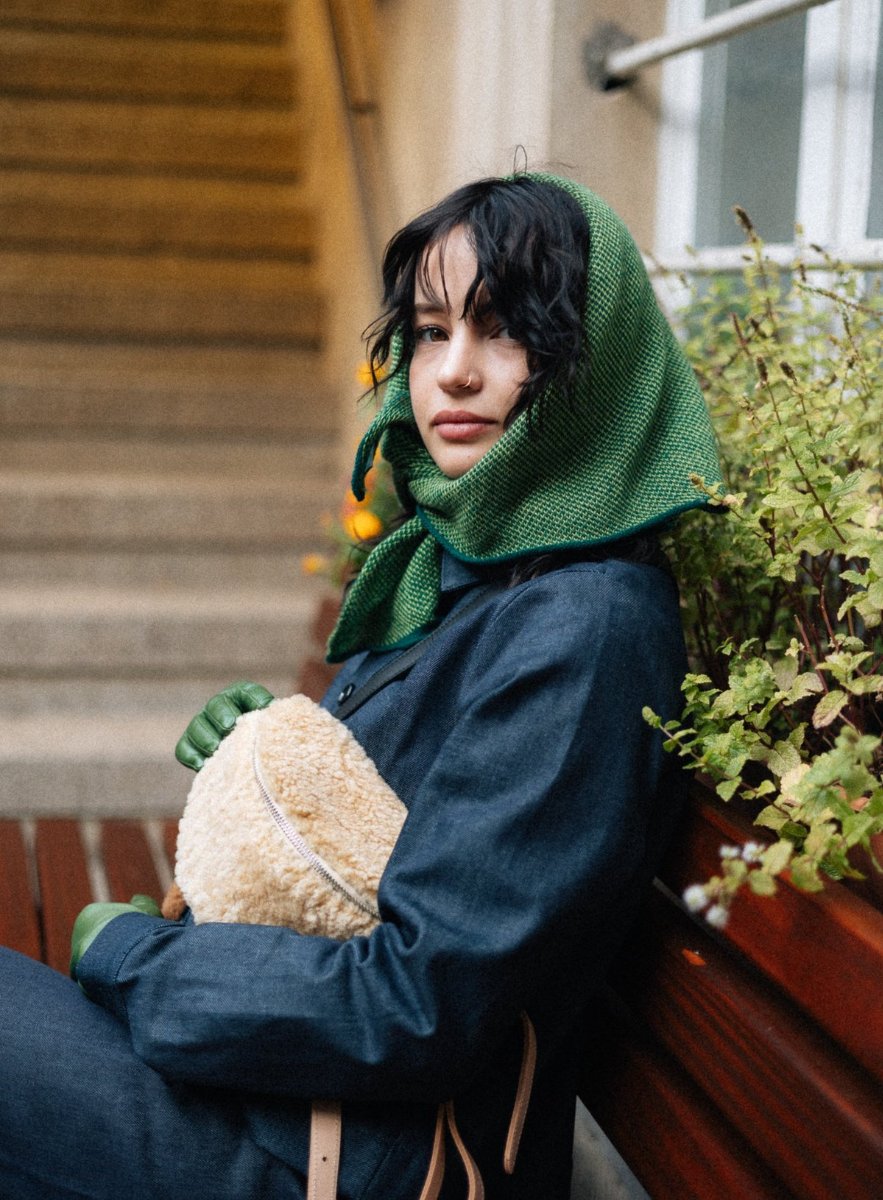 A woman in a green headscarf on a wooden bench holds a cream shearling hip bag to her chest.