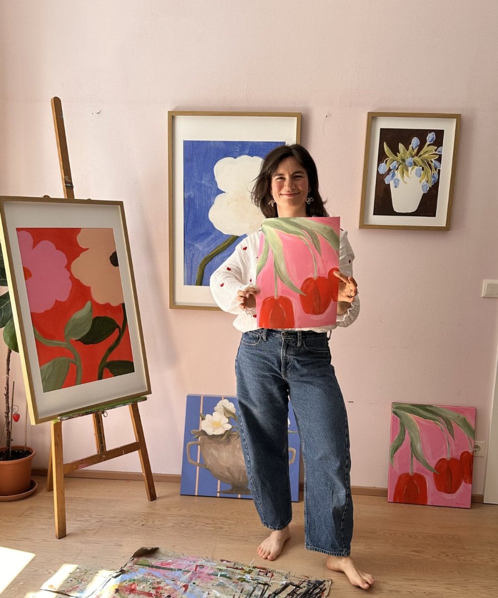 Johanna Blaha stands barefoot in her studio holding one of her paintings, an easel and more of her work behind her.