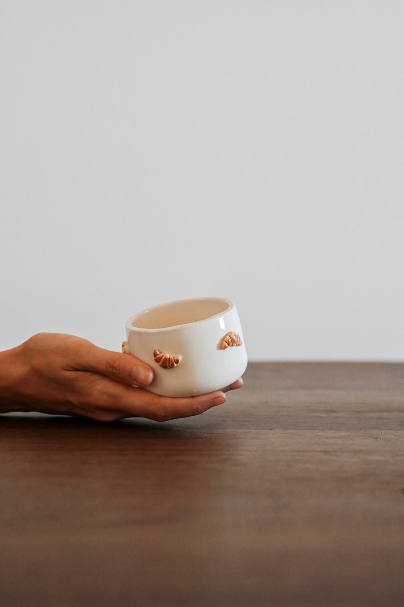 A hand lifting a cup with a small croissant stuck to its side