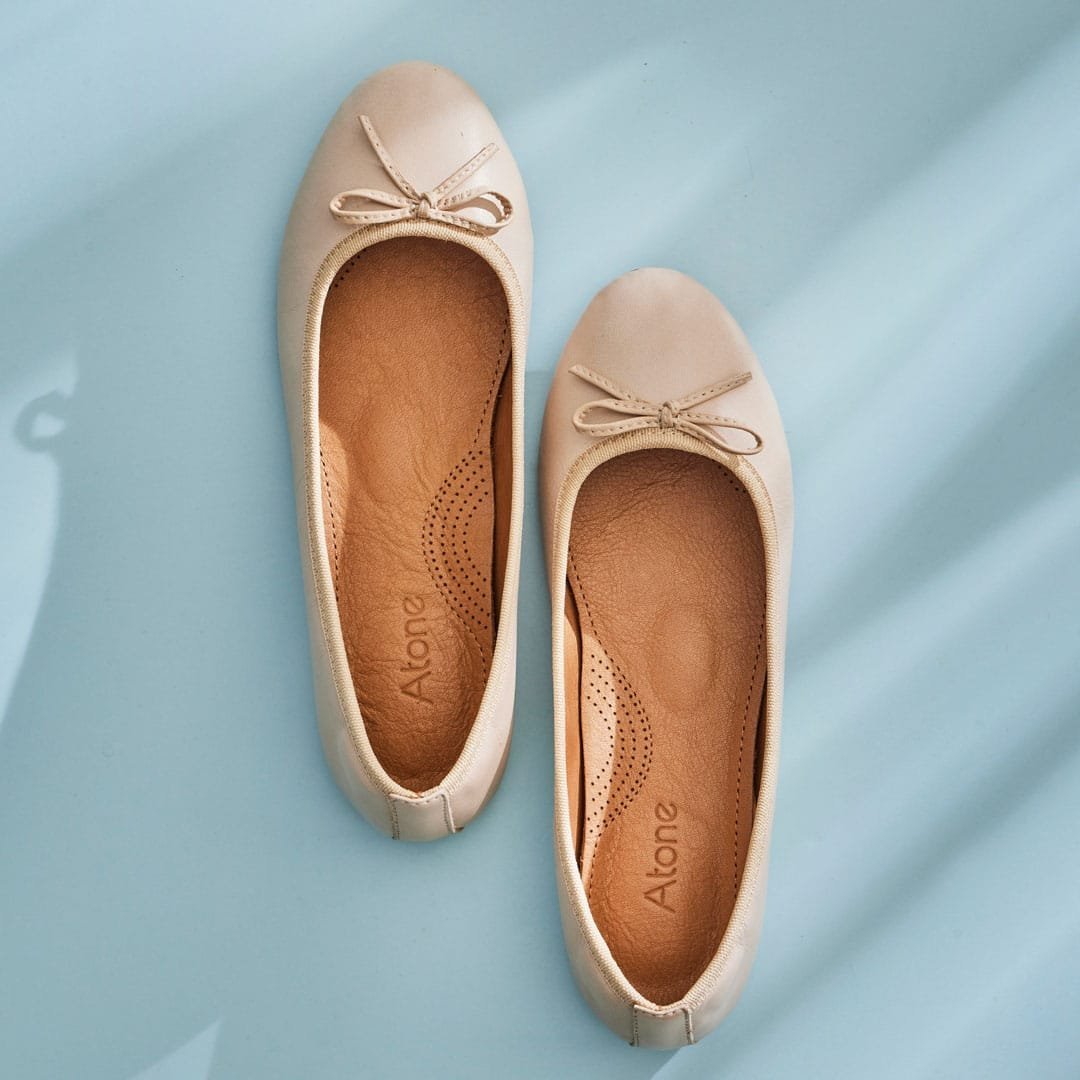 Two cream ballerina flats with small bows and Atone wordmarks on the tan insoles lie diagonally on a pale blue surface.