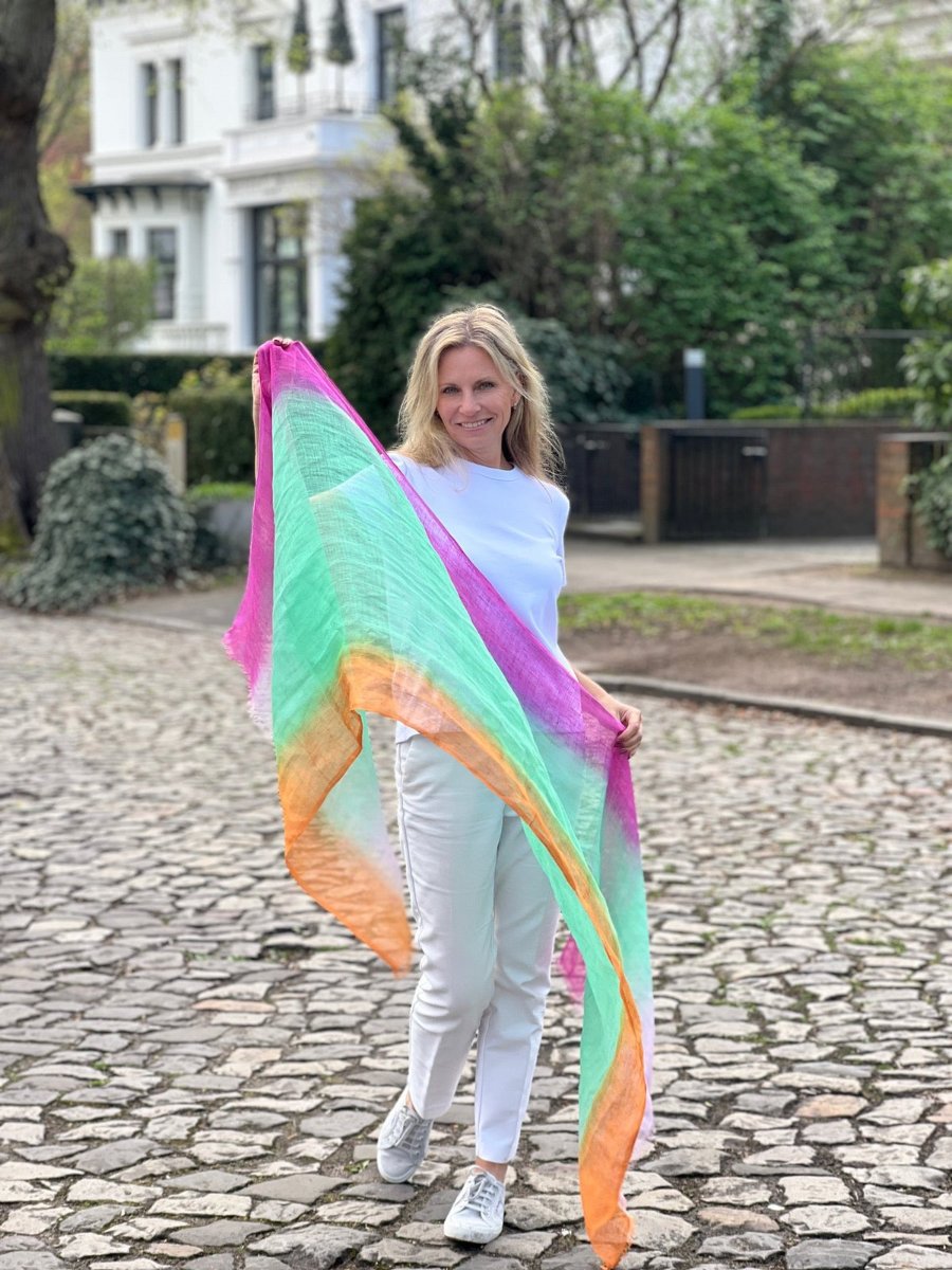 A woman on a cobbled street holds a large pink, mint and orange scarf into the wind