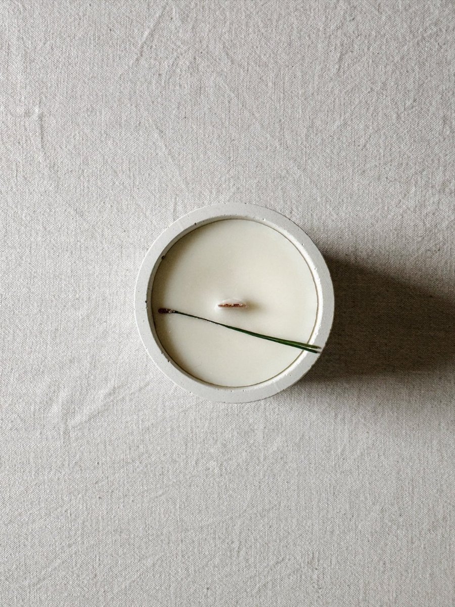 A concrete vessel filled with soy and rapeseed wax, the wooden wick lying flat across the middle, two spruce needles on top.
