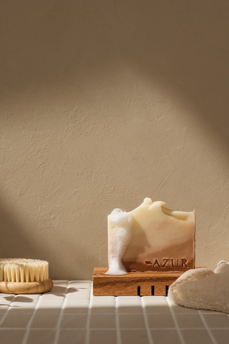 A bar left standing on a slatted wooden dish on a tiled bathroom ledge, a wooden brush and a konjac sponge beside it.