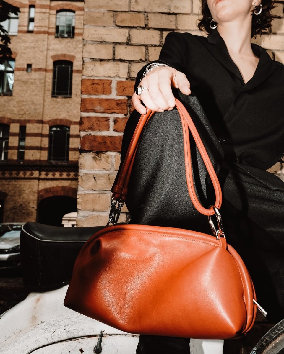 The Loulou baguette in cognac, held up by its handle against a brick façade.