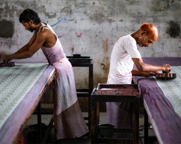 Two printers at a long padded table, the cloth pulled out flat, blocks in hand.