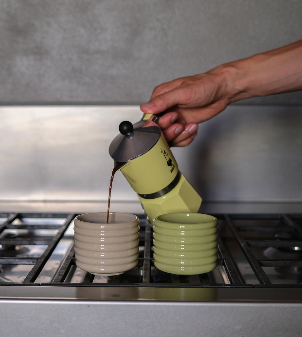 A hand pours coffee from a moka pot into a cup on the hob.