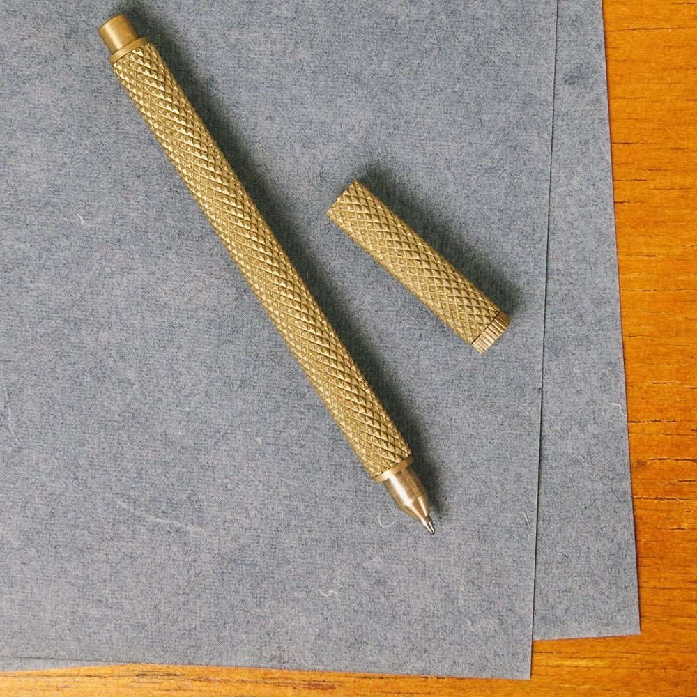 The knurled brass pen on grey paper, its cap set down beside it.
