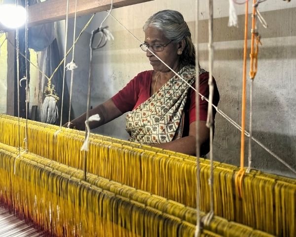 A weaver at a pit loom, the yellow warp stretched away from her under tension.