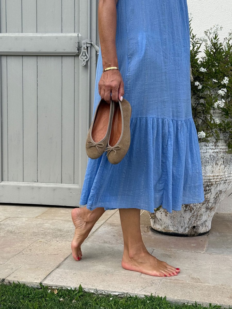 Daylight, grass and a pale wooden door. A woman walks barefoot, a pair of brown flats carried in one hand.